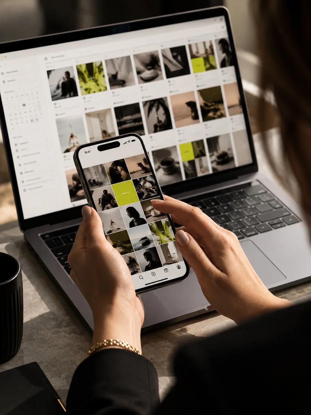 Hands holding a phone showing an Instagram feed, with the content calendar open on a laptop behind