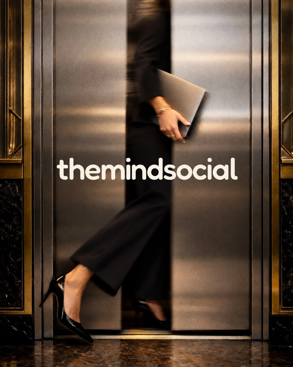 A woman in black carrying a laptop walks past brass elevator doors, the themindsocial wordmark across the frame